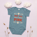 Teal baby onesie with 'Not All Girls Wear Pink' text on a light pink background with white flowers and dried plants.