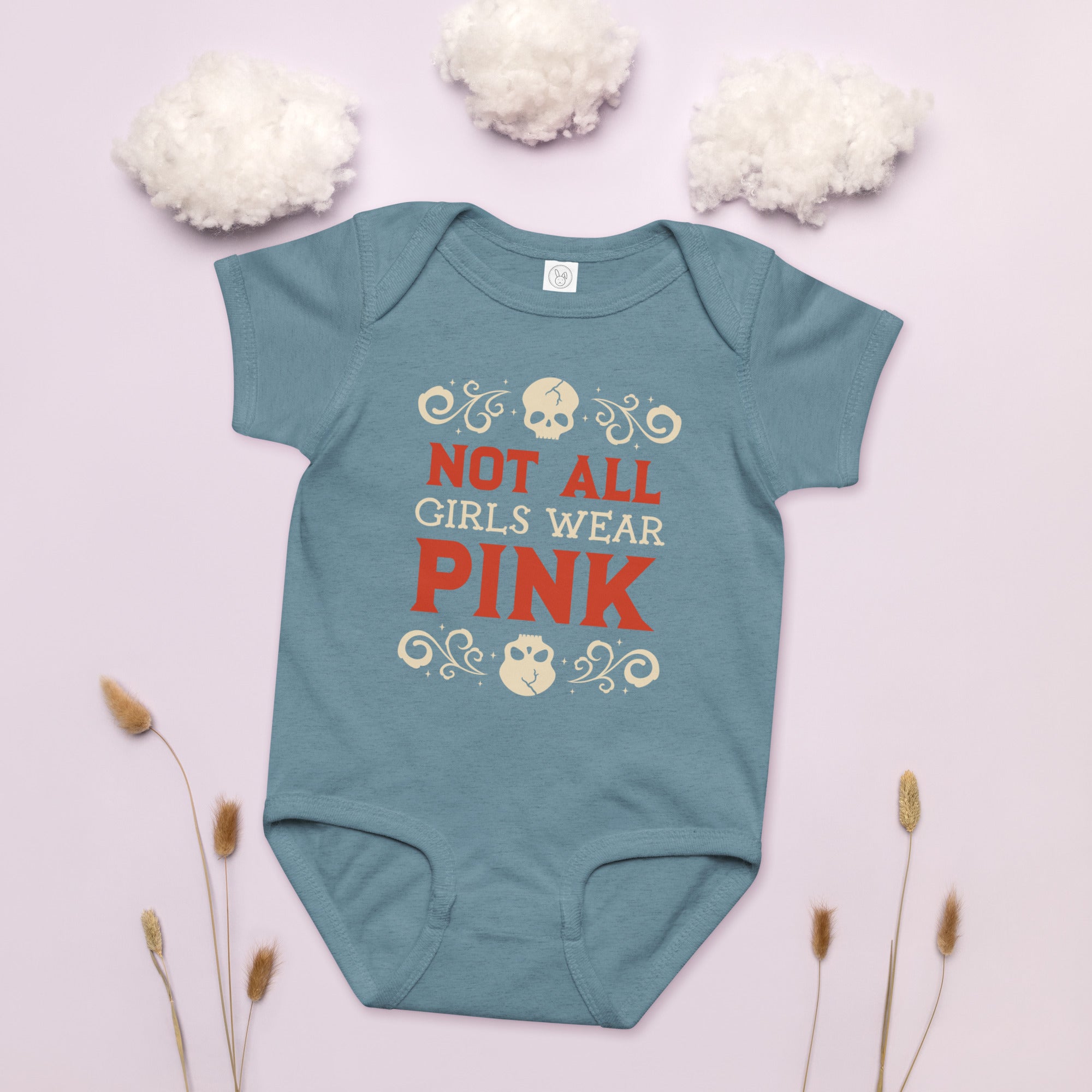 Teal baby onesie with 'Not All Girls Wear Pink' text on a light pink background with white flowers and dried plants.