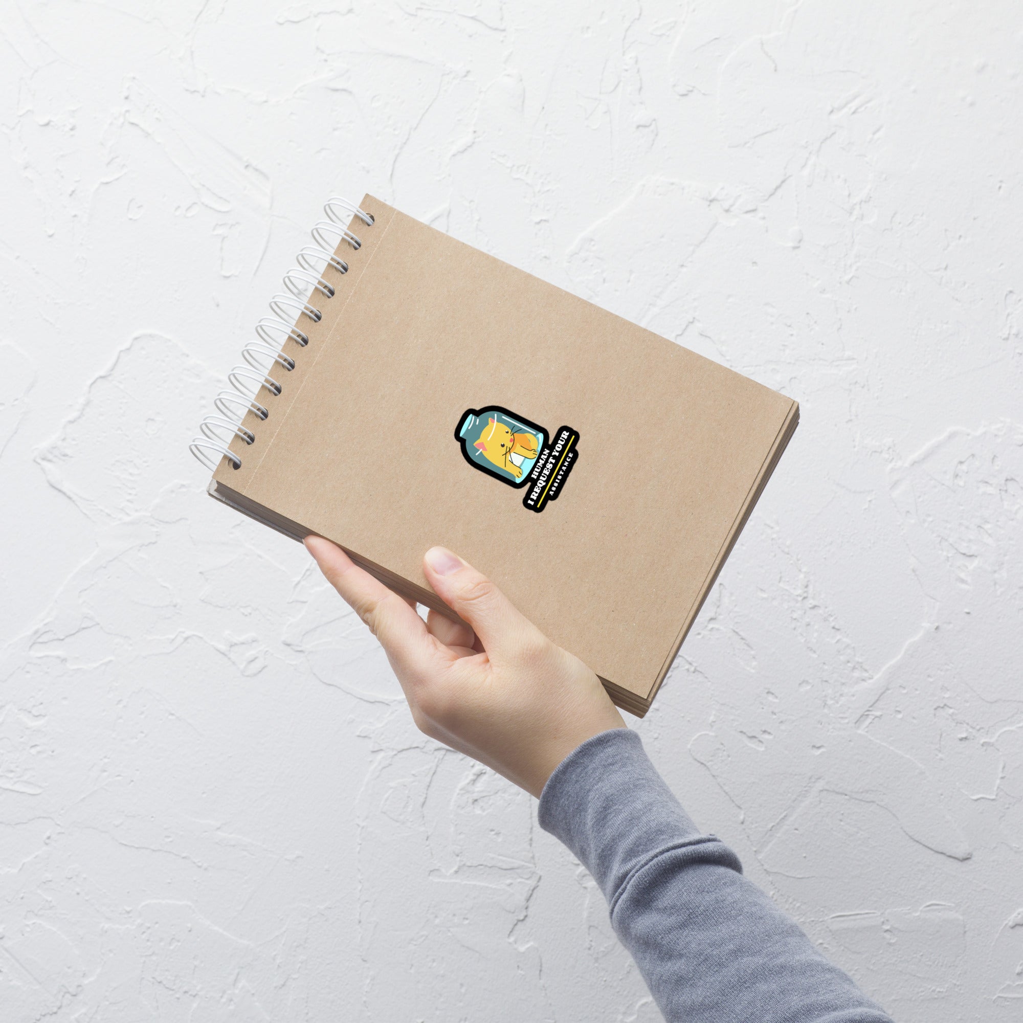 Hand holding a brown notebook with a sticker on a light gray textured background