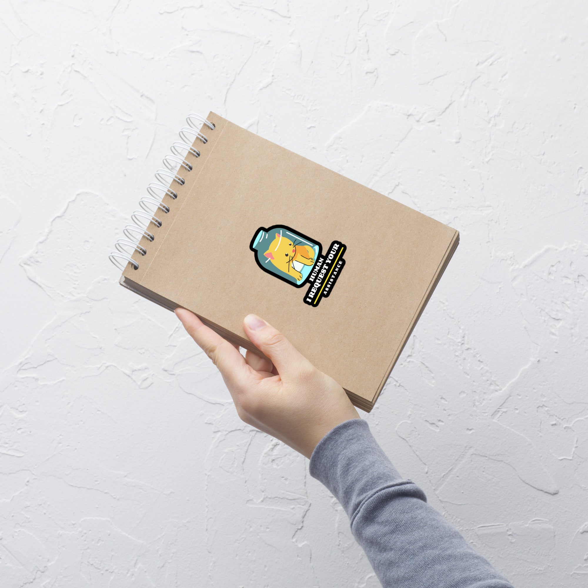 Hand holding a brown notebook with a sticker on a light gray textured background