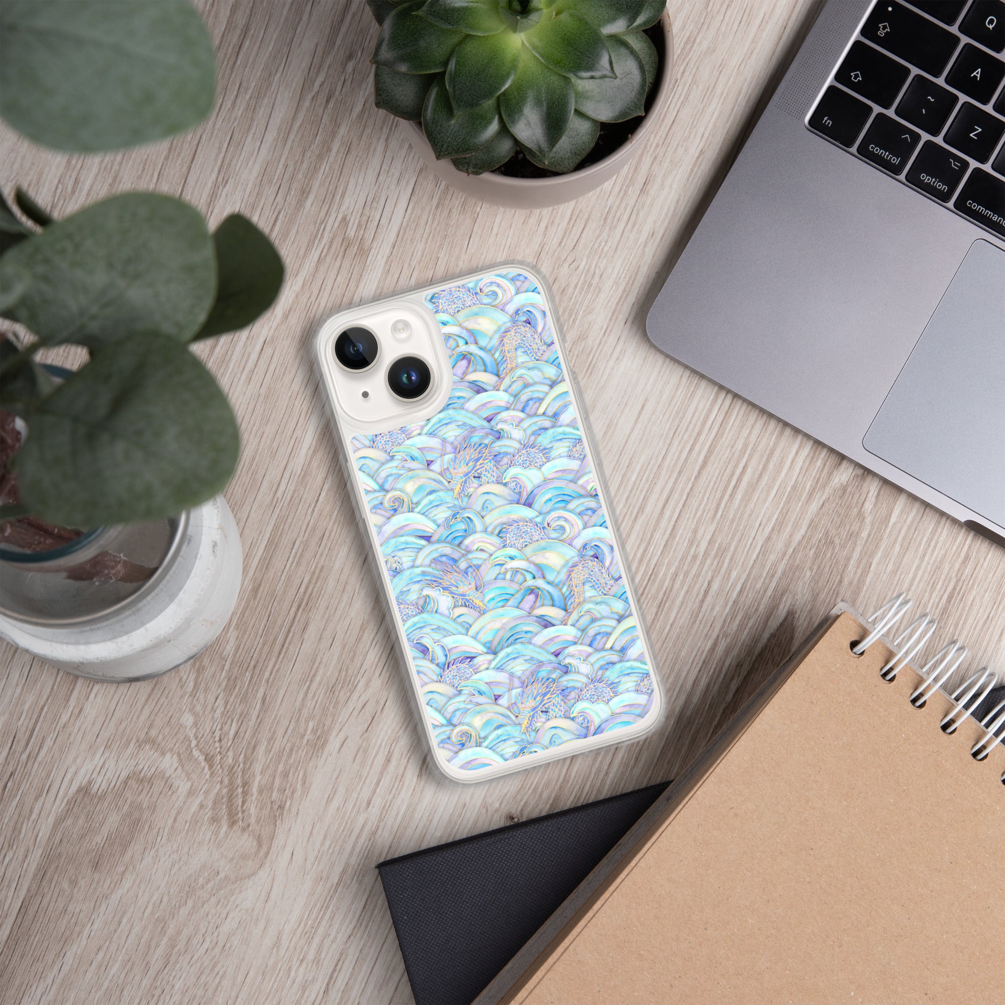 Phone with a colorful case on a desk next to a laptop, plant, and notebook.