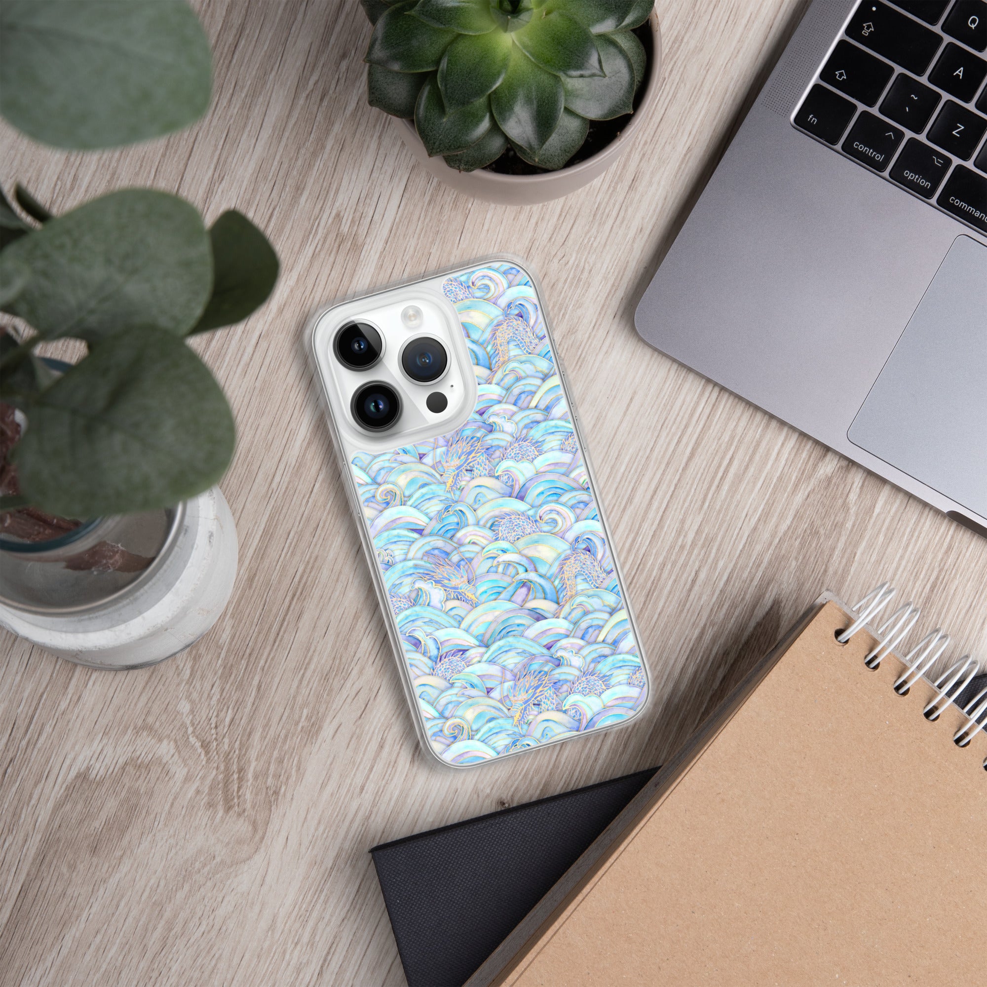 Phone with a colorful case on a desk next to a laptop, plant, and notebook.