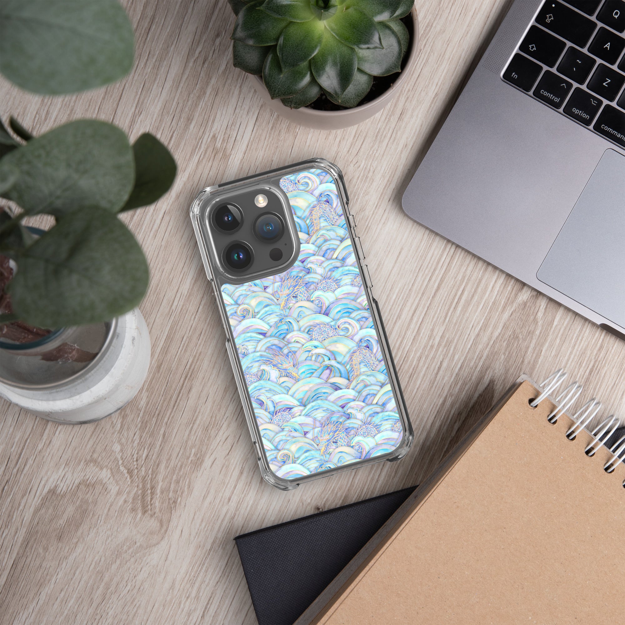 Phone case with a colorful pattern on a desk with a laptop, notebook, and plants.