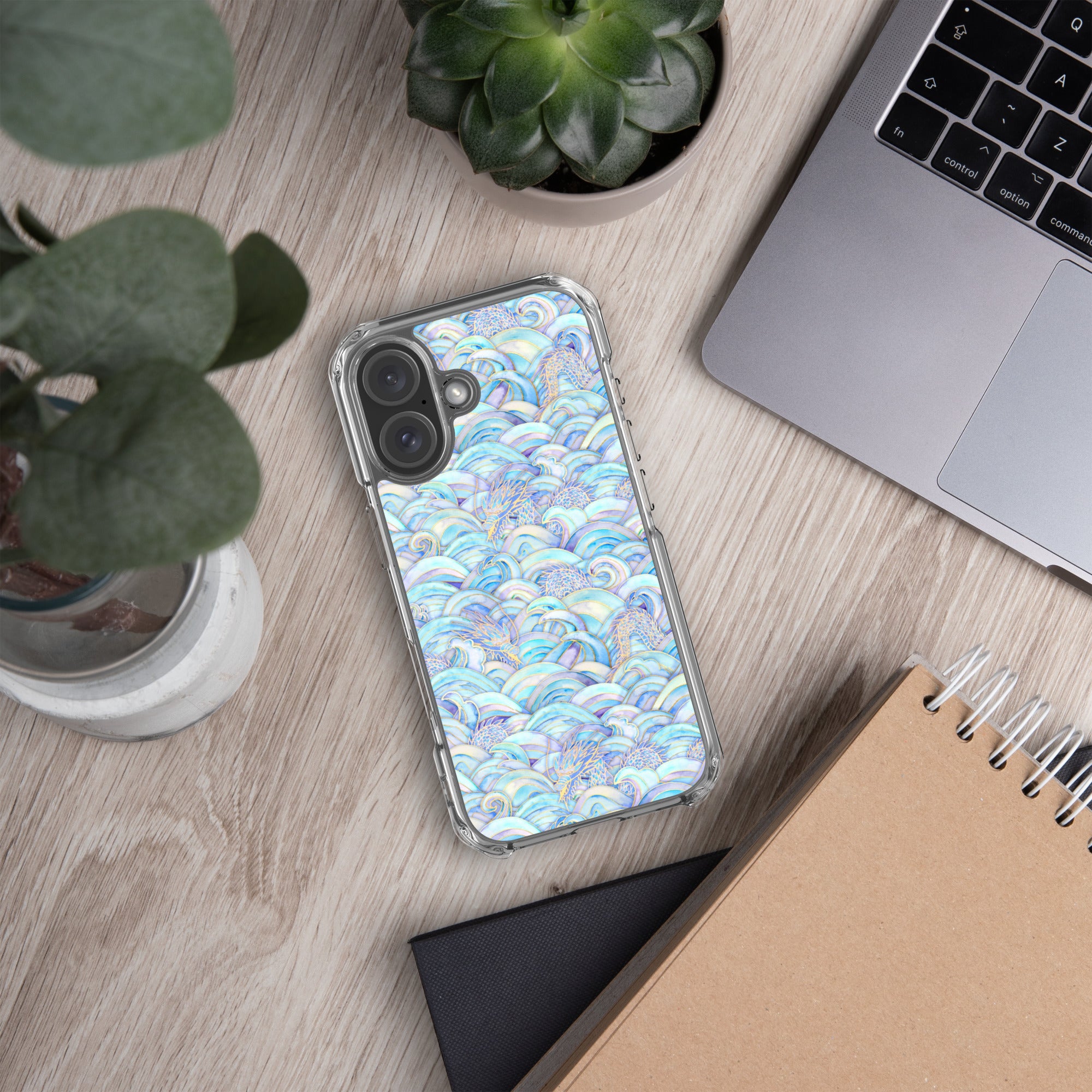 Phone with a colorful case on a desk next to a laptop, plant, and notebook.