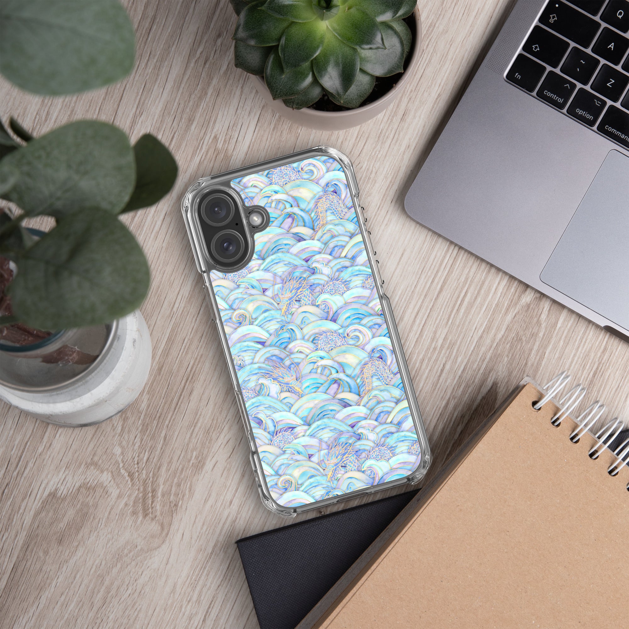 Phone case with blue and white pattern on a wooden desk with laptop, notebook, and plant.